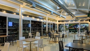 A spacious indoor dining area with multiple tables and chairs, large windows offering city views, and visible air ducts on the ceiling.