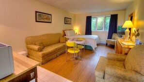 A cozy room with a bed, a sofa, a small table, and a chair. There are two lamps on the table and a window with curtains. - Picket Fence Motel (St. Andrews)