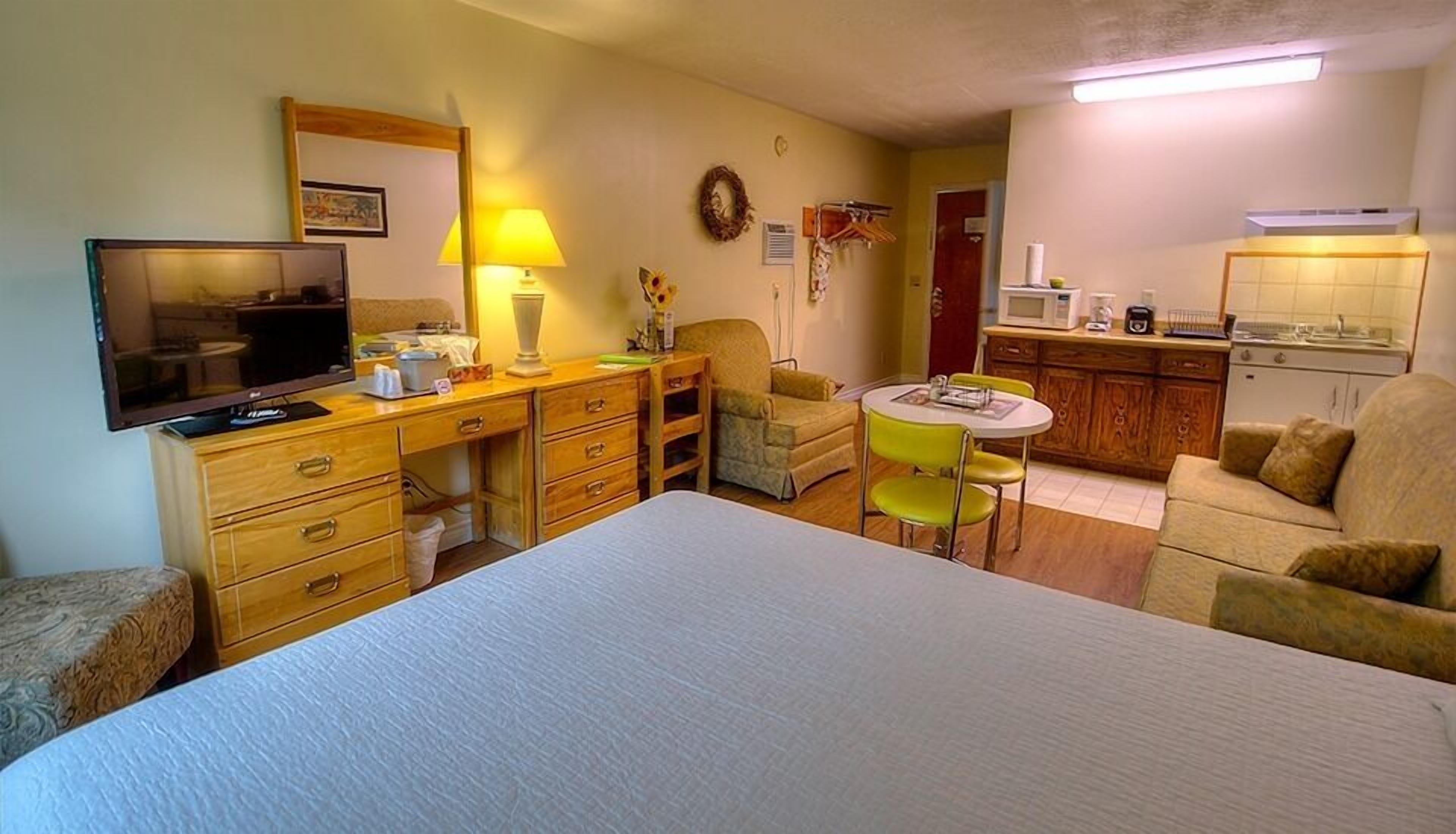 A compact hotel room with a bed, a desk, a chair, a small dining table, a microwave, and a kitchenette.