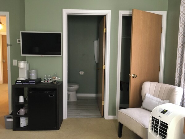A hotel room with a television, a small refrigerator, a seating area with a chair, and a door leading to a bathroom.