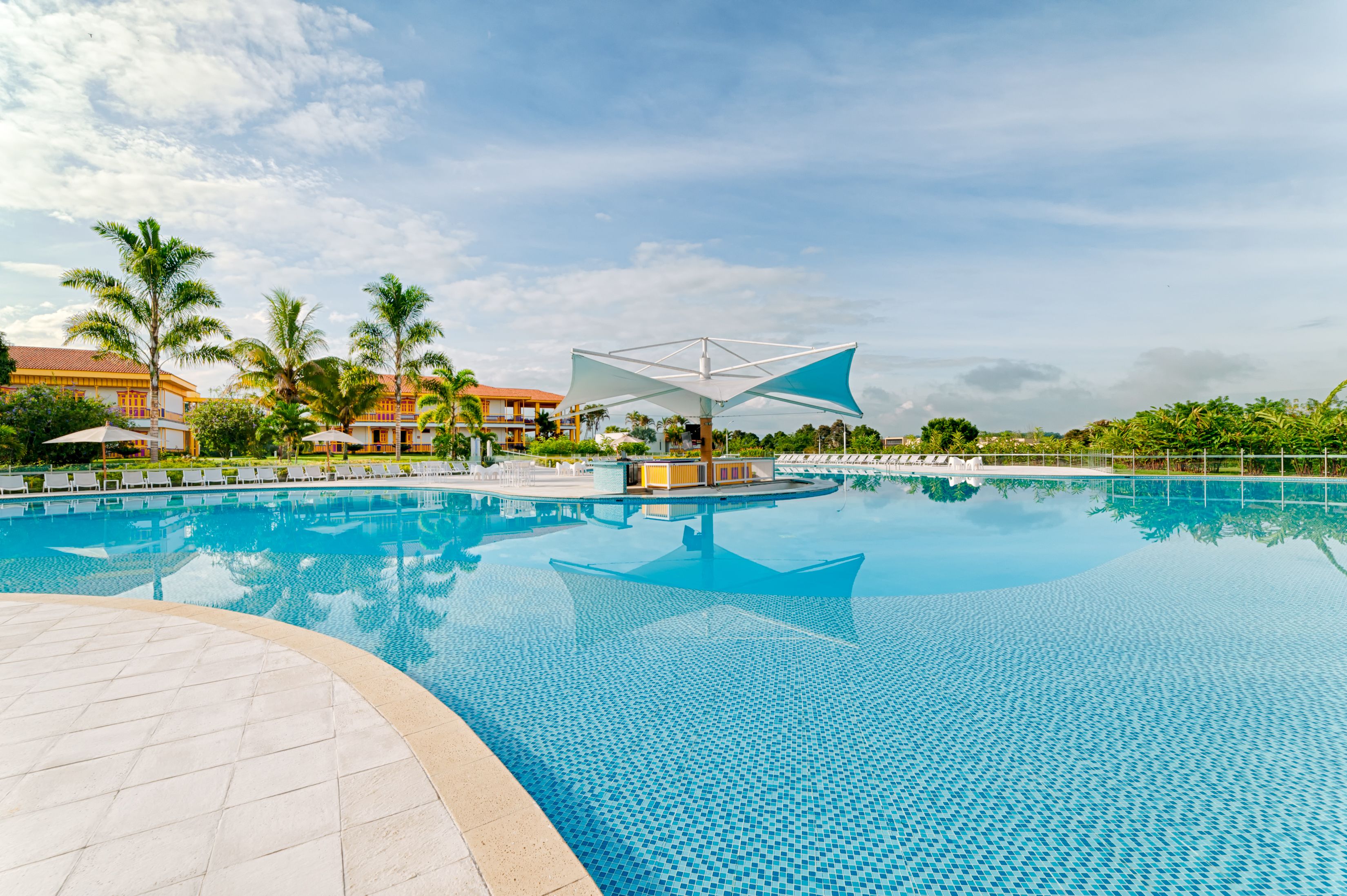 A large swimming pool with a central sunshade, surrounded by palm trees and lounge chairs.