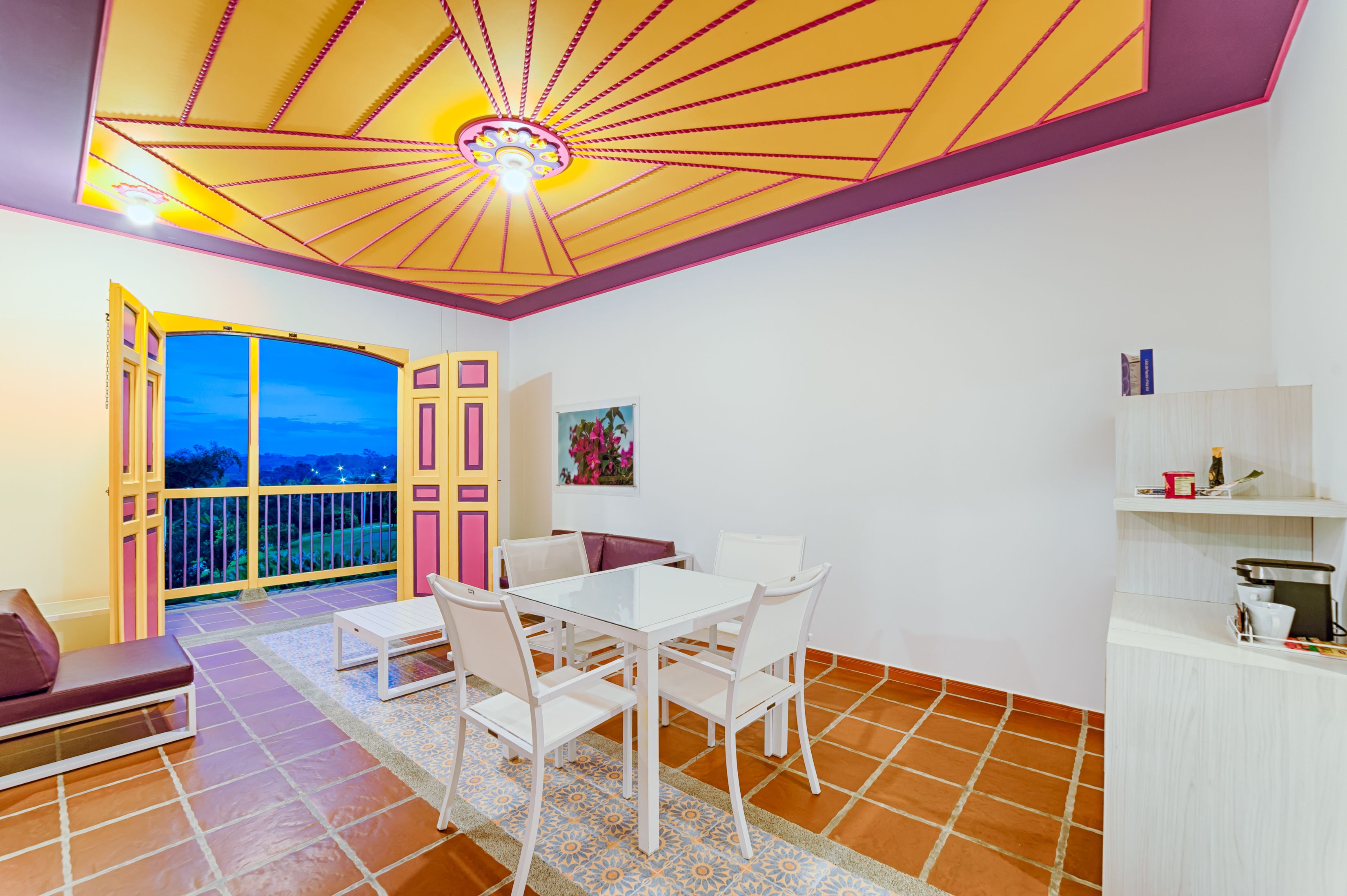 A dining area with a white table and chairs, a red cushioned bench, and a view through an open door to the outside.