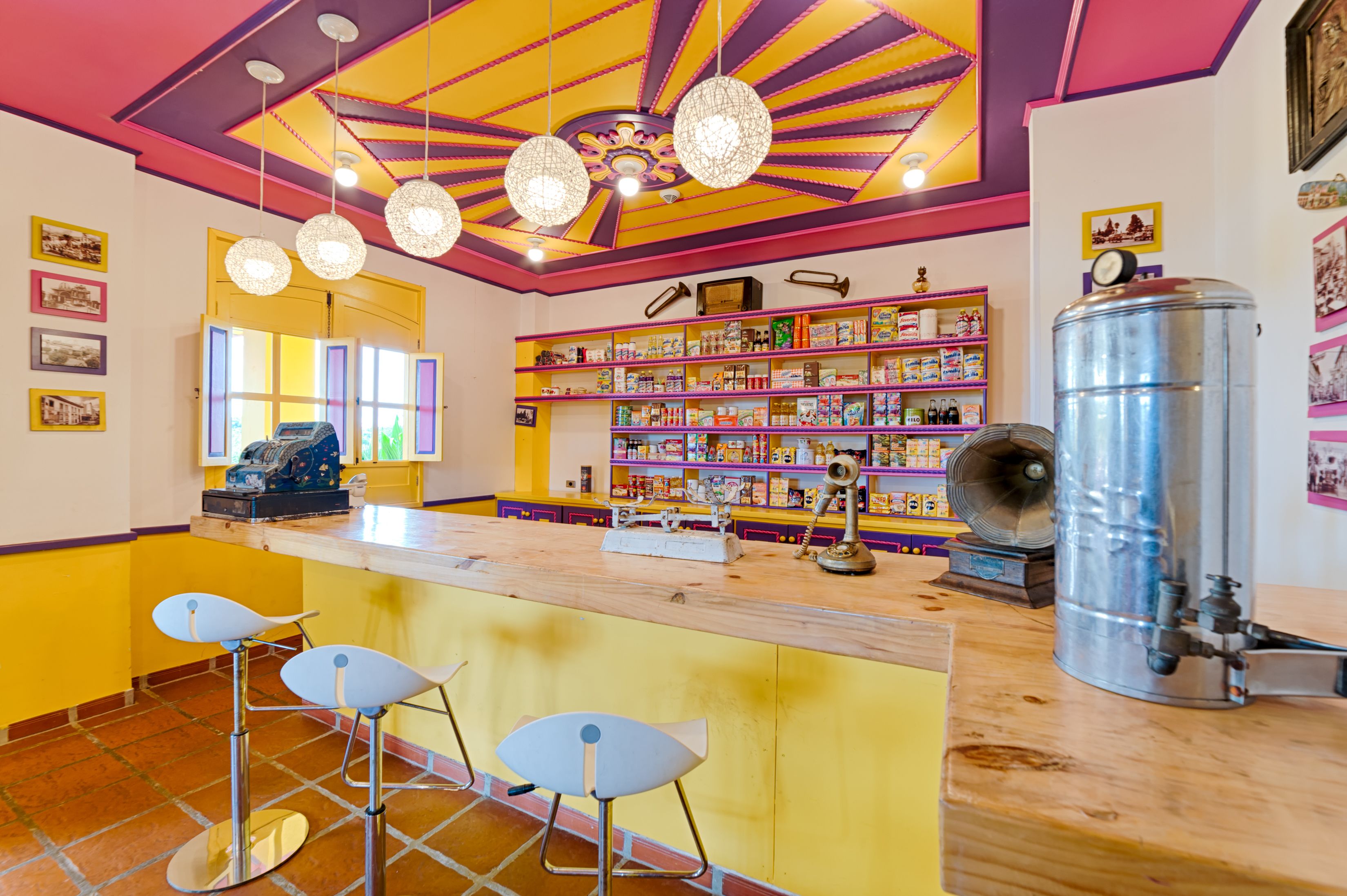 A brightly colored bar area with a wooden counter, bar stools, and shelves stocked with various items.