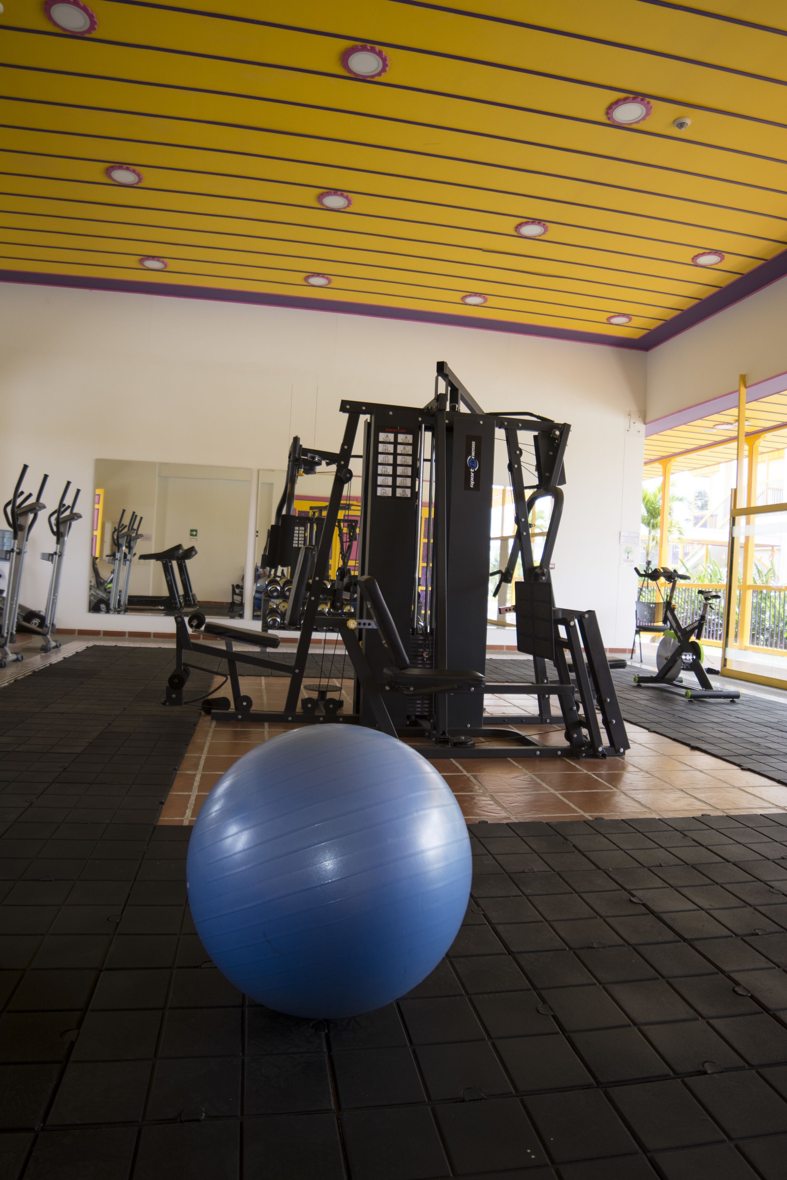 Un gimnasio con equipamiento de ejercicio, una pelota azul para ejercicio y un techo amarillo.