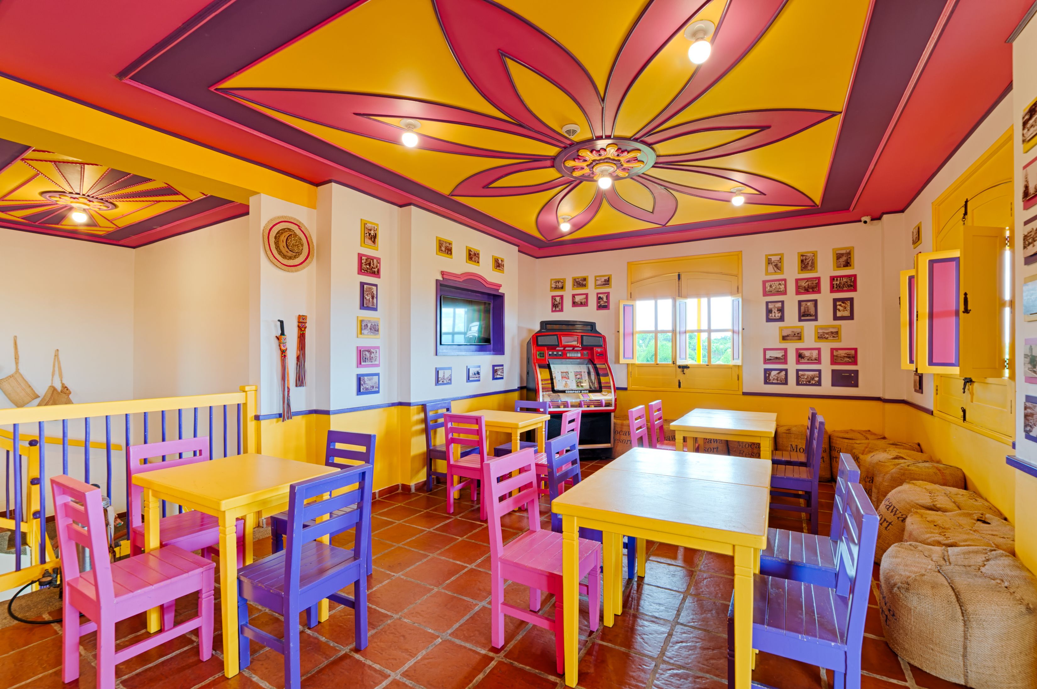 Un restaurante de colores vivos con mesas y sillas amarillas y rosas, un techo con diseño floral y un radiolas.
