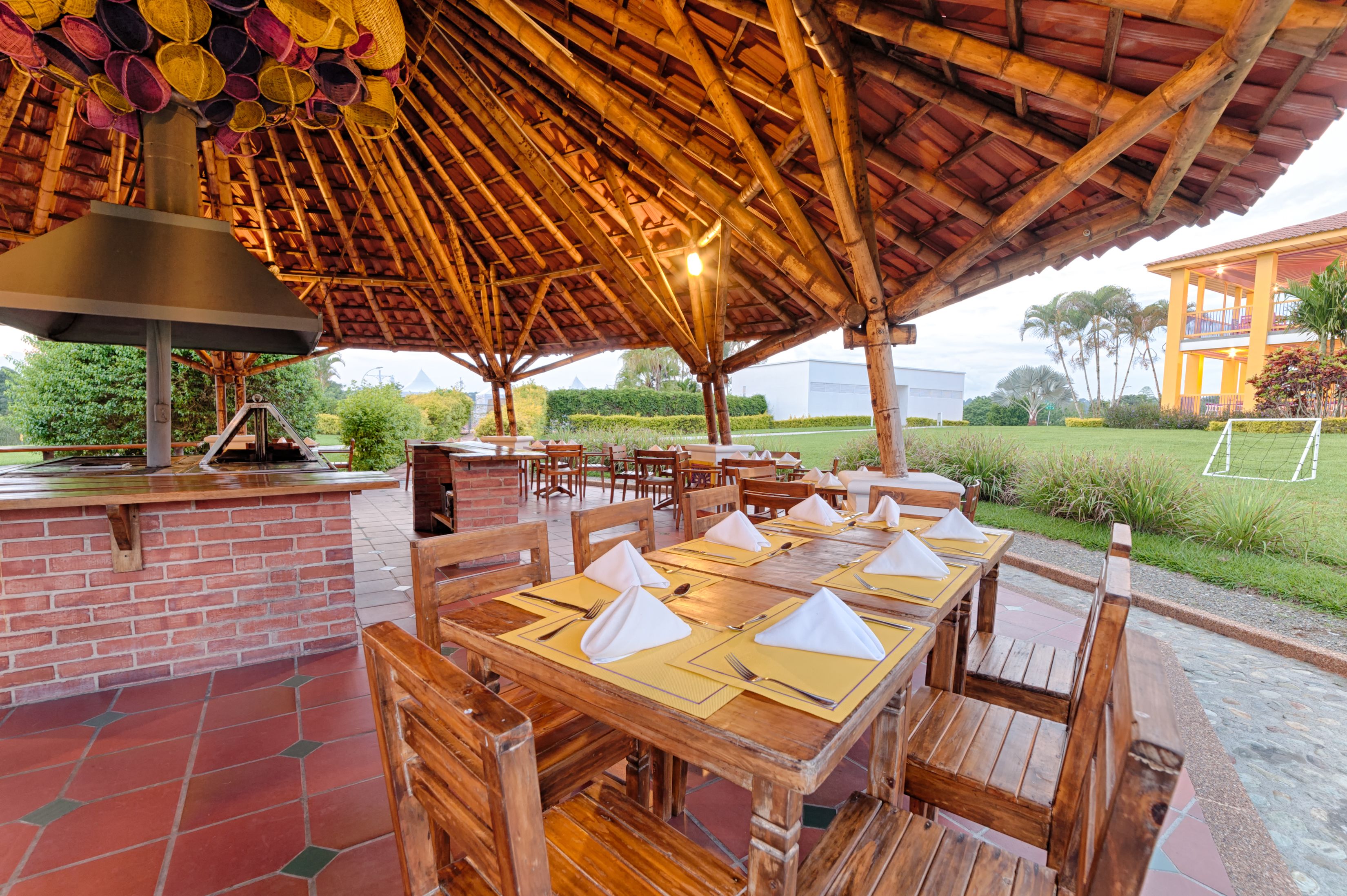 Un espace extérieur aménagé pour les repas, avec des tables et des chaises en bois, un barbecue en briques et une structure dotée d’un toit de chaume.