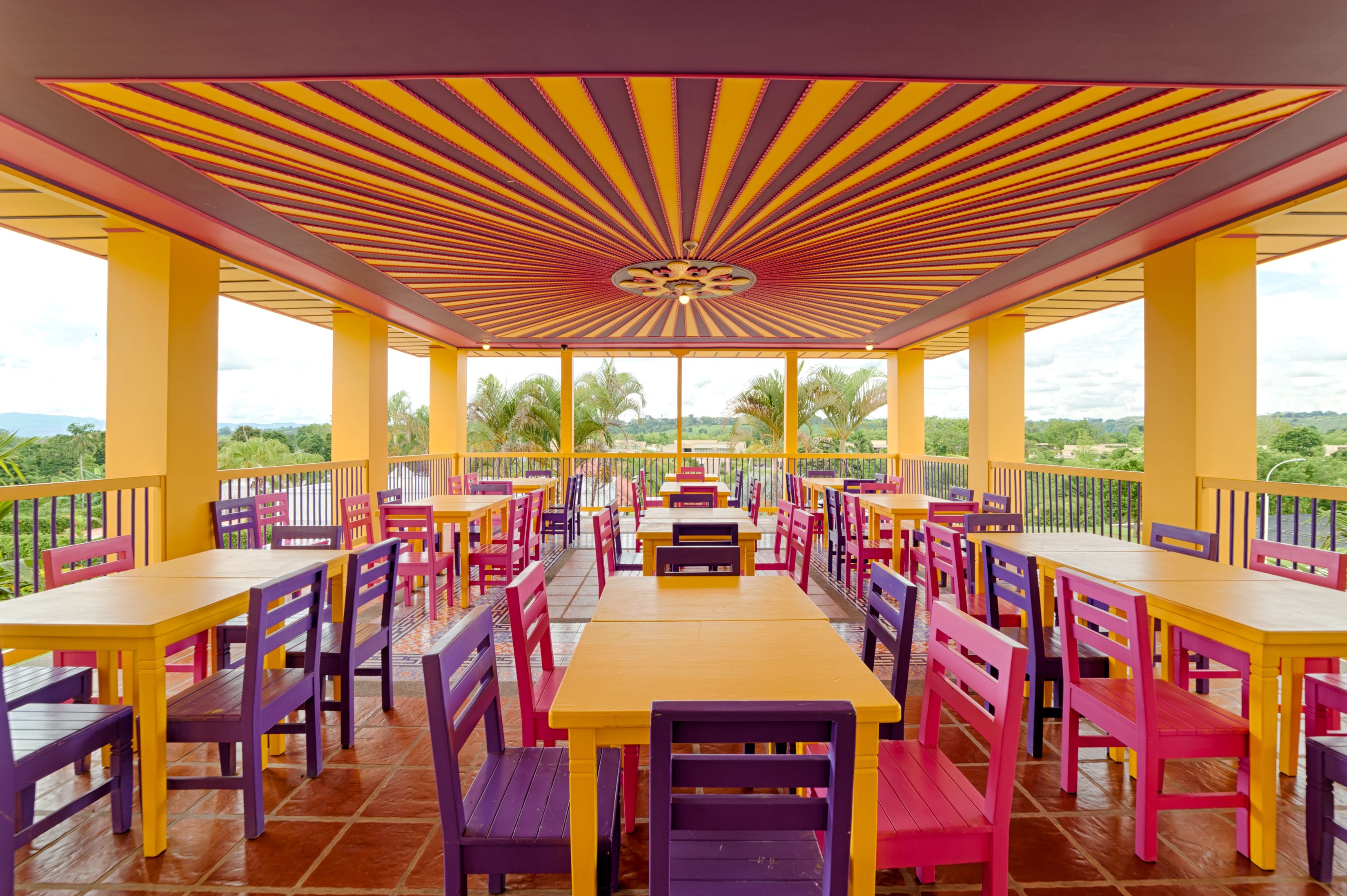 Un espace extérieur abrité, aménagé pour les repas, avec des tables et des chaises jaunes et violettes, un sol carrelé et une vue sur la verdure et les collines en arrière-plan.