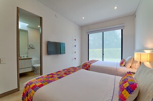 Quarto de hotel com duas camas, televisão, janela com persianas e banheiro visível através de uma porta aberta.