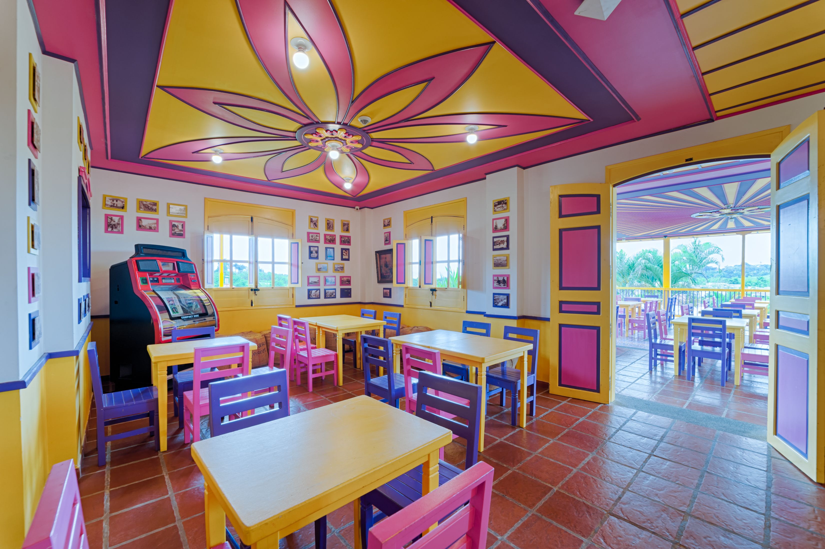 Un restaurant aux couleurs vives, avec des tables et des chaises jaunes et roses, un plafond orné de motifs floraux et une vue sur l’extérieur grâce à de grandes fenêtres.