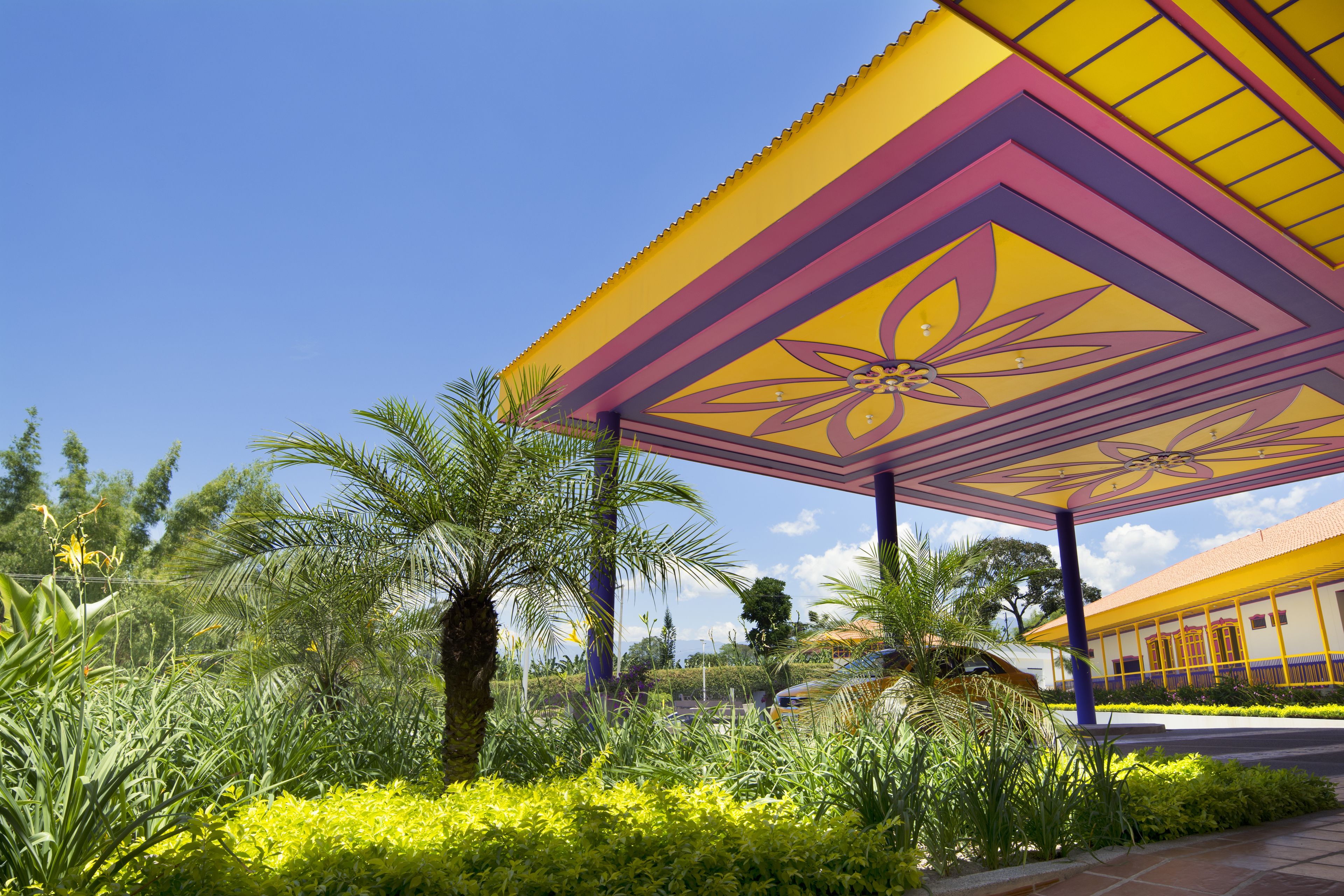 A covered outdoor area with a geometric roof design, surrounded by lush greenery and palm trees.