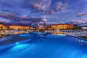 A large outdoor swimming pool with a modern architectural structure in the center, surrounded by well-lit buildings and lounge areas. - Hotel Mocawa Resort (La Tebaida)