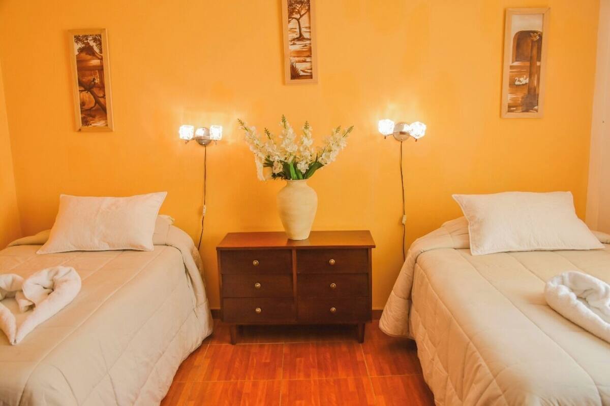 Habitación de hotel con dos camas, una mesita de noche con un florero y luces fijadas a la pared.