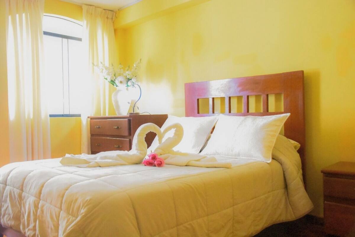 Una cama bien hecha con edredón blanco y almohadas, cabecera de madera, mesita de noche con un florero y ventana con cortinas.