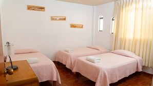 A hotel room with three beds, each with pink bedding and white towels. There are two framed pictures on the wall, a window with curtains, and a wooden desk with a lamp. - Hospedaje Claudia (Ica)