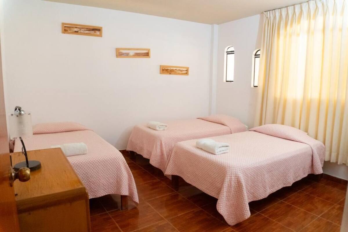 Habitación de hotel con tres camas, cada una con ropa de cama rosa y toallas blancas. Hay dos cuadros en la pared, una ventana con cortinas y un escritorio de madera con una lámpara.