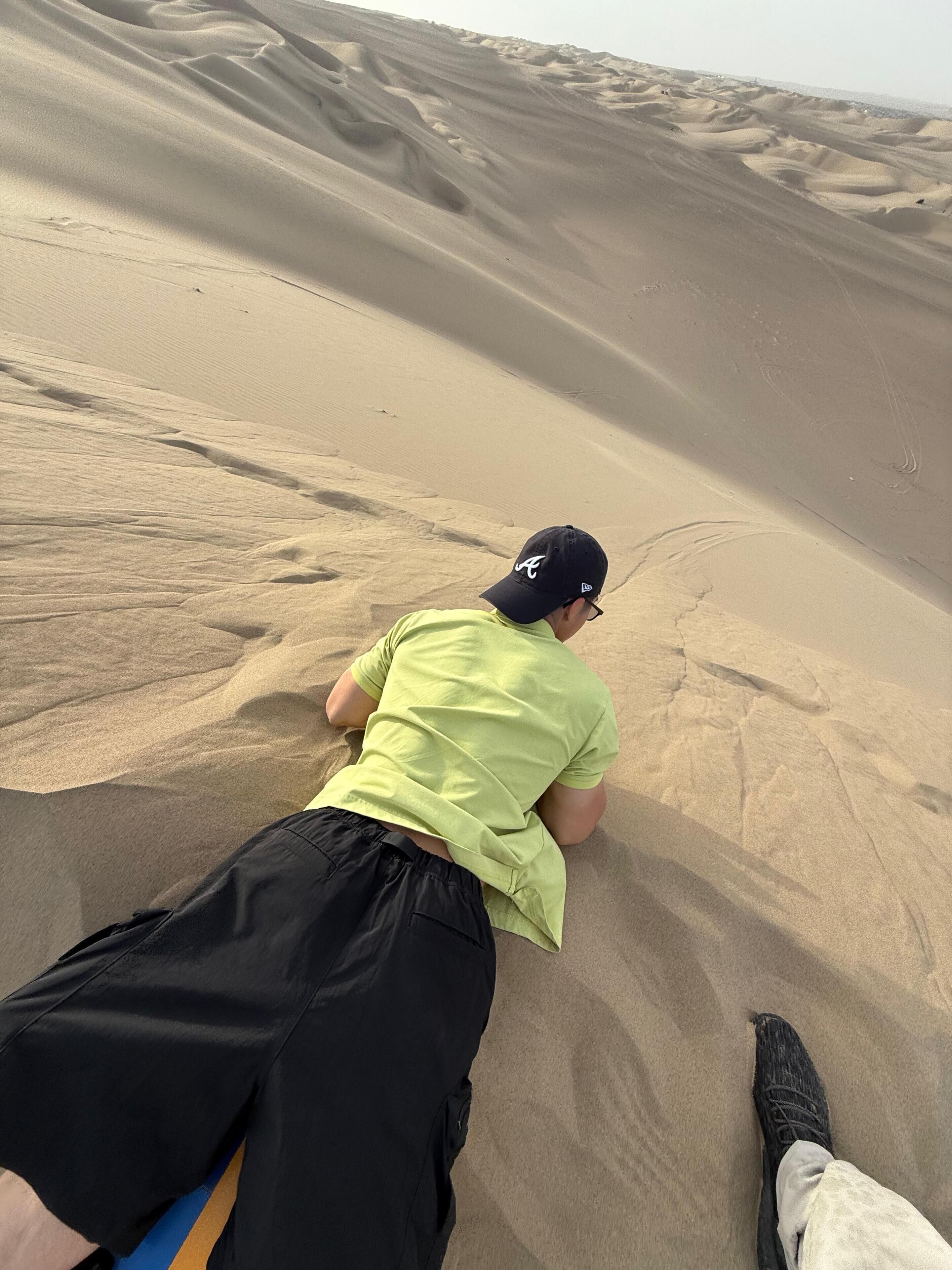 A person lying on sand dunes, wearing a cap and a green shirt.