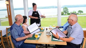 A dining area with a table set for four people, a waiter standing by, and a view of a lake through large windows. - Cabot Trail Motel (Baddeck)