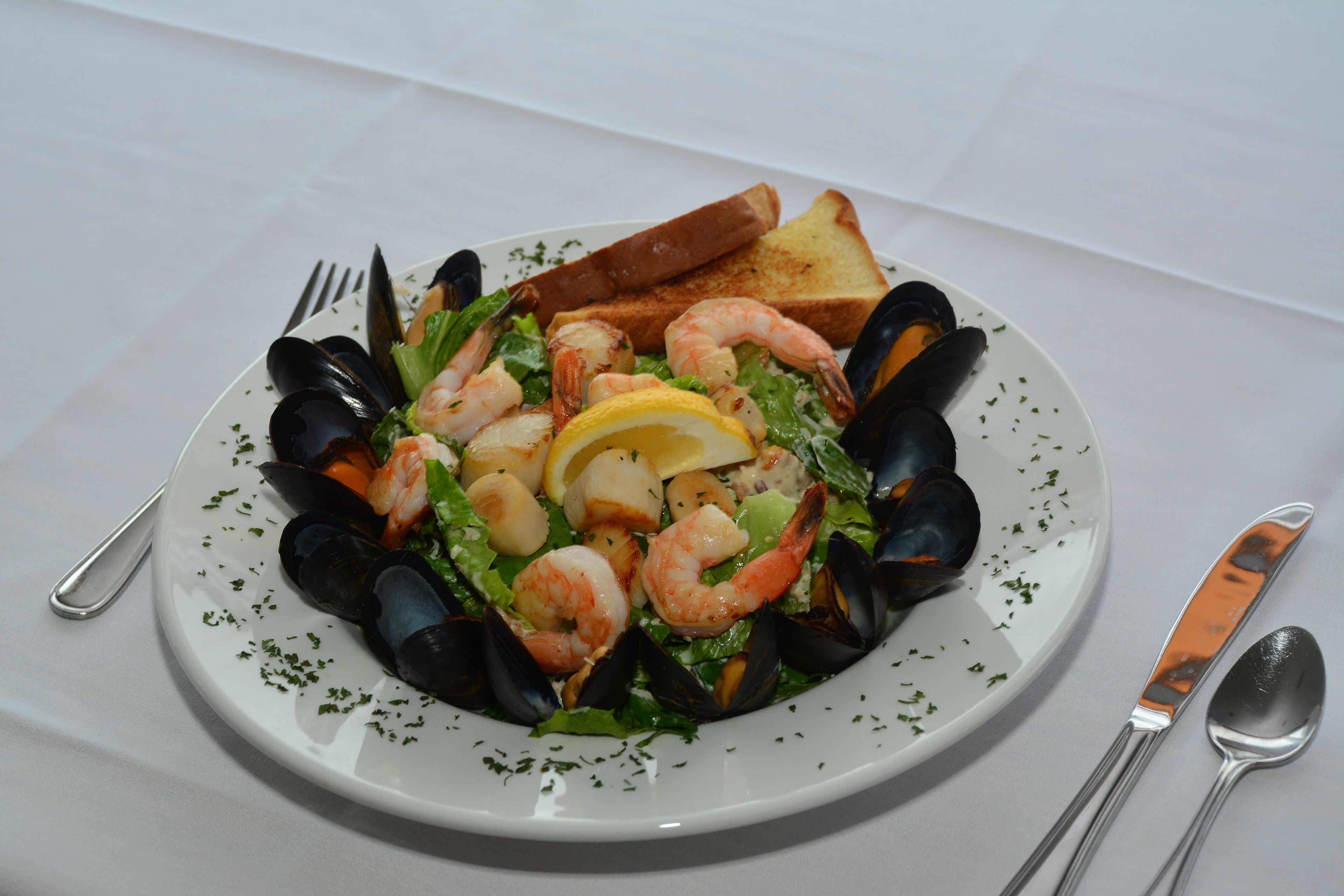 Un plato de mariscos con mejillones, camarones y una cuña de limón, acompañado de tostadas y cubiertos.