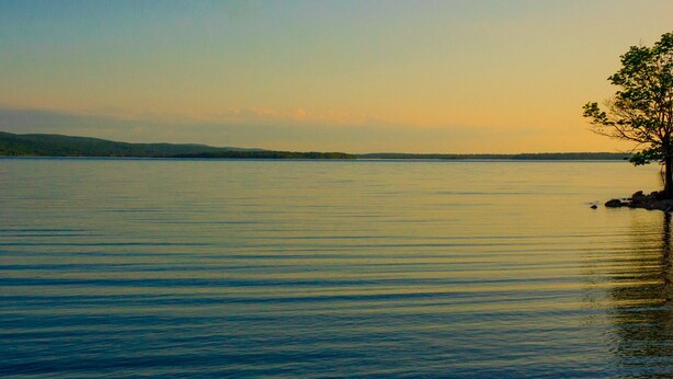 A calm lake with a tree on the right and hills in the background. - Cabot Trail Motel (Baddeck)