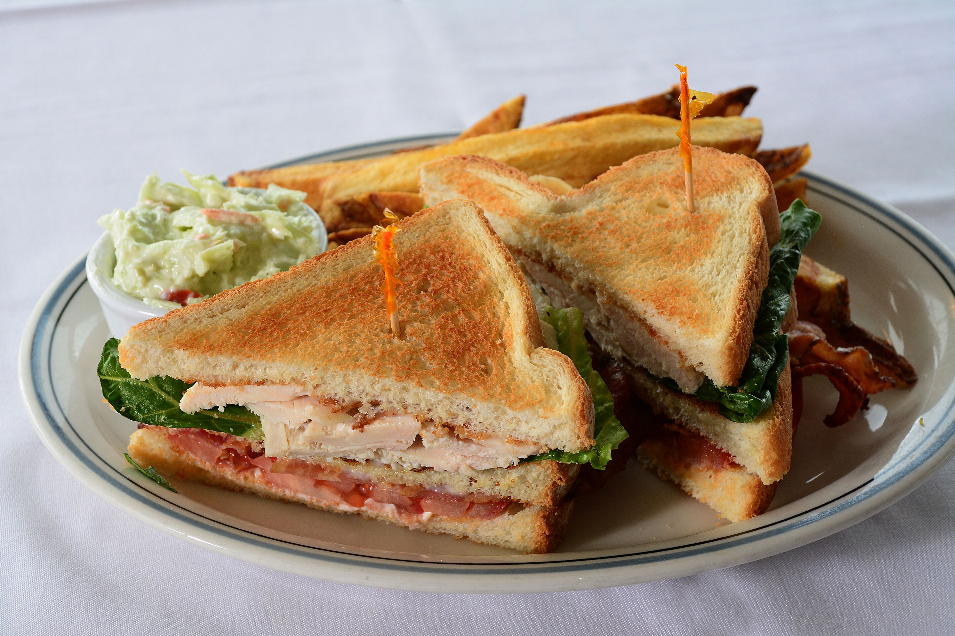 A sandwich cut in half with bacon, lettuce, and tomato, served with a side of coleslaw and fries on a plate.