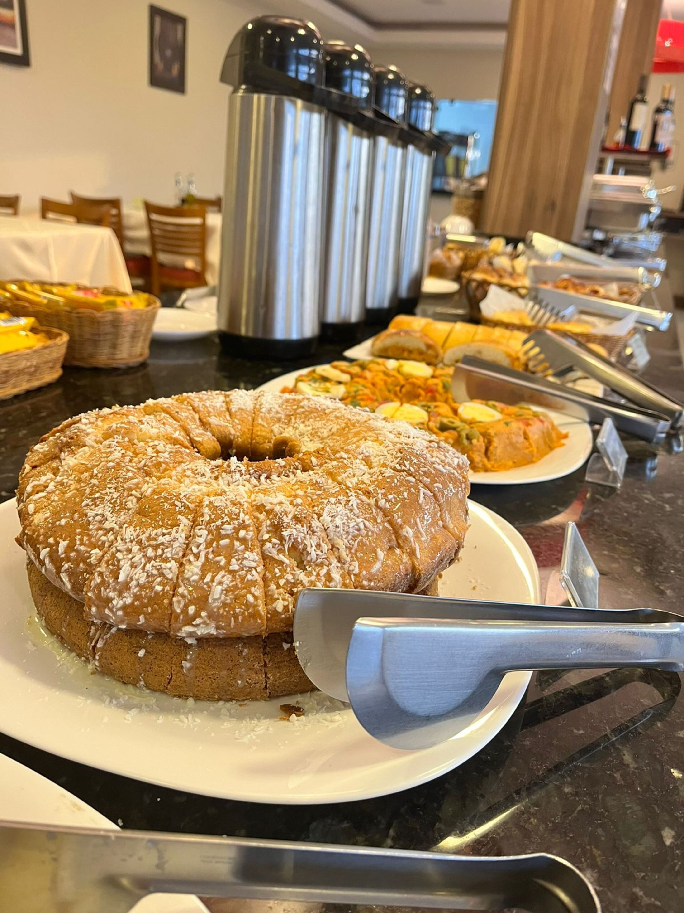 Buffet com diversos pratos, incluindo um bolo grande com coco ralado e um espaço para café com vários potes térmicos.