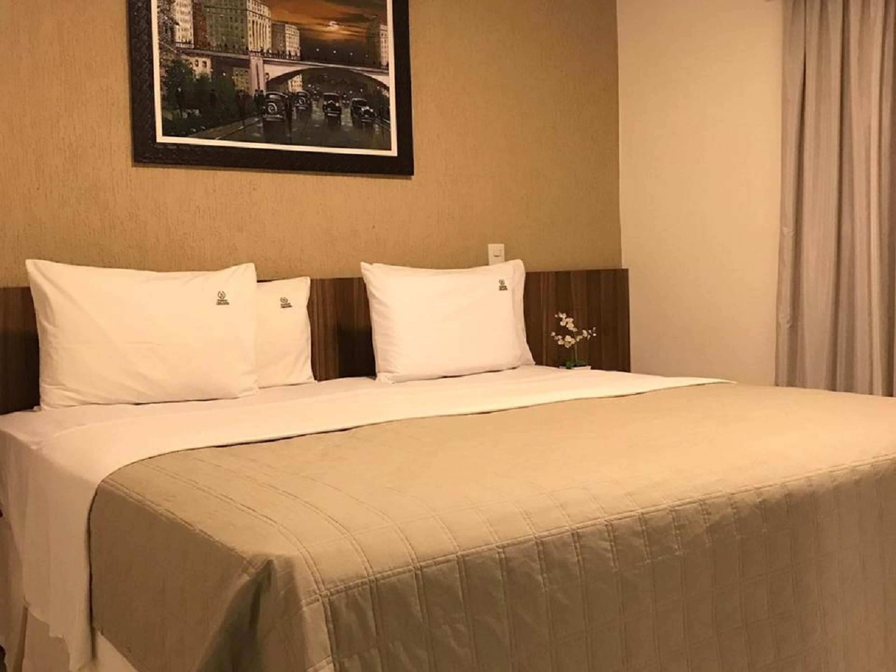 A hotel room with a bed, white pillows, a beige bedspread, a framed picture on the wall, and a nightstand with a vase of flowers.