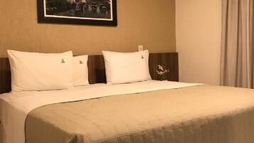 A hotel room with a bed, white pillows, a beige bedspread, a framed picture on the wall, and a nightstand with a vase of flowers.