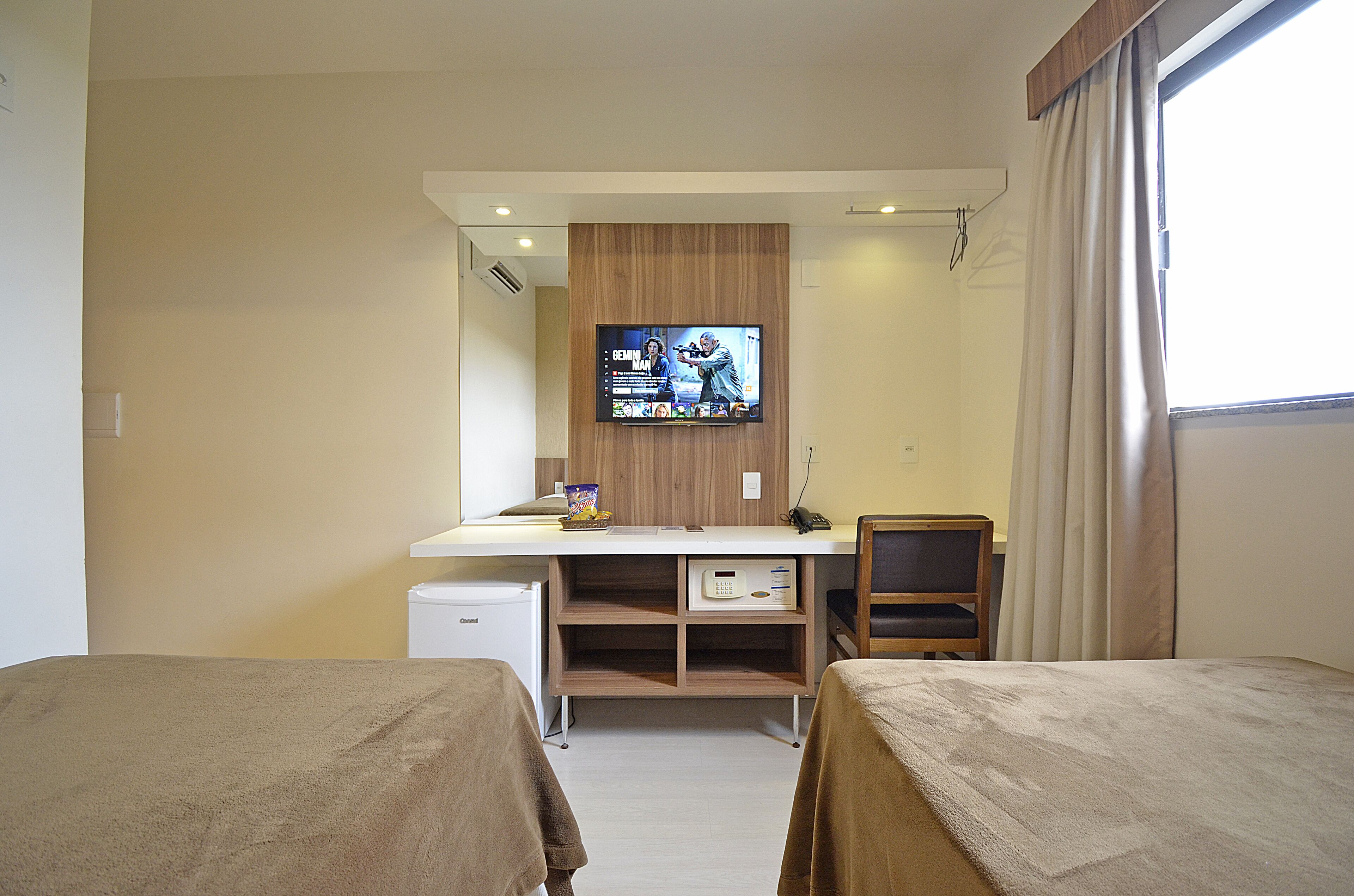 Una habitación de hotel con cama, escritorio con televisor, refrigerador pequeño y ventana con cortinas.
