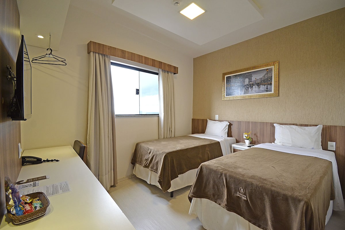 A hotel room with two beds, a desk with a telephone and a basket of snacks, a window with curtains, and a framed picture on the wall.