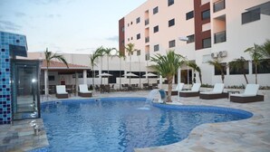 Ein Swimmingpool mit Liegestühlen und Sonnenschirmen, umgeben von einem Gebäude mit mehreren Etagen und Balkonen.