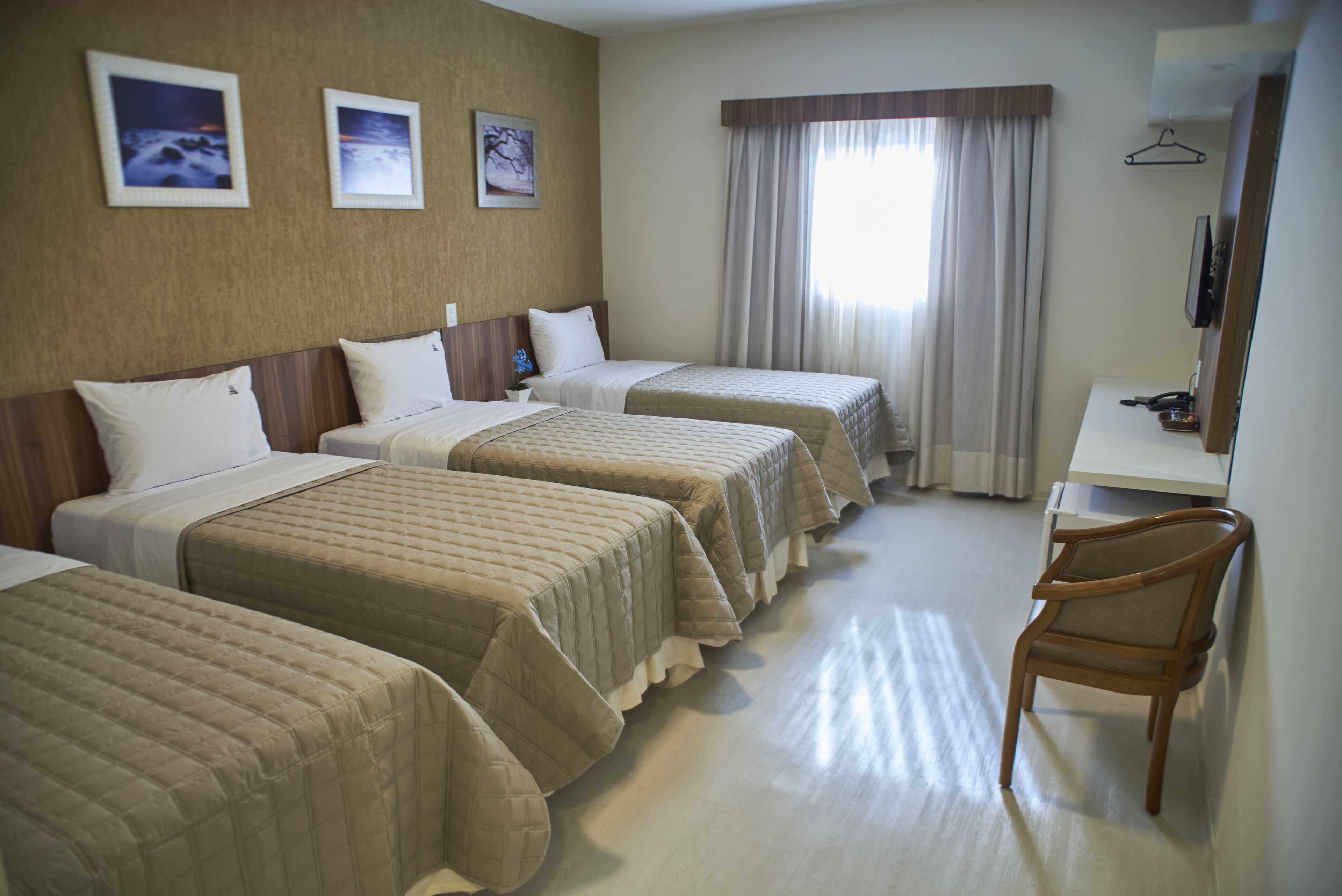 A hotel room with three beds, a desk, and a chair. There are three framed pictures on the wall.