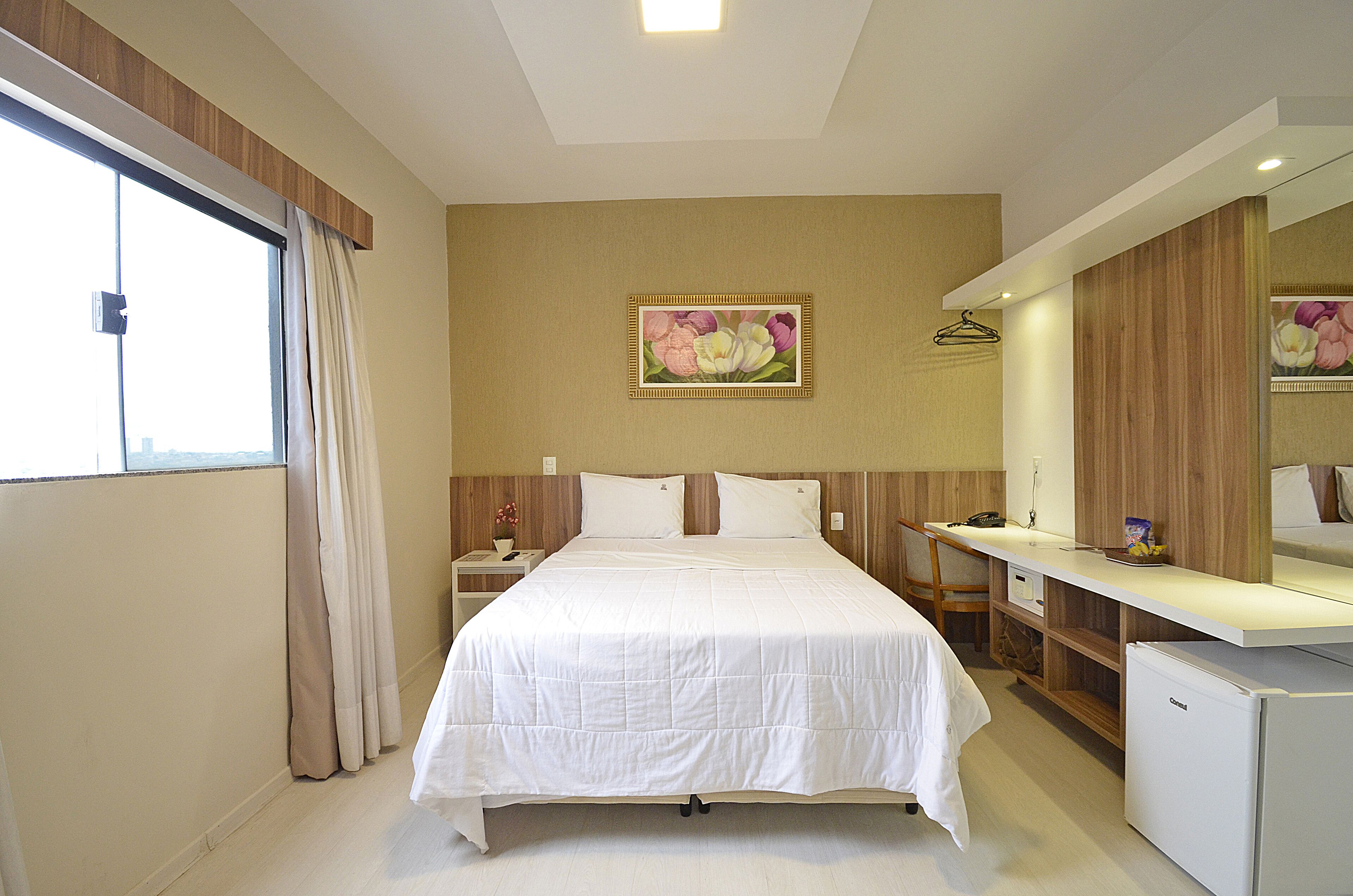 Habitación de hotel con cama, escritorio, refrigerador, mesita de noche, ventana con cortinas y un cuadro con motivos florales en la pared.