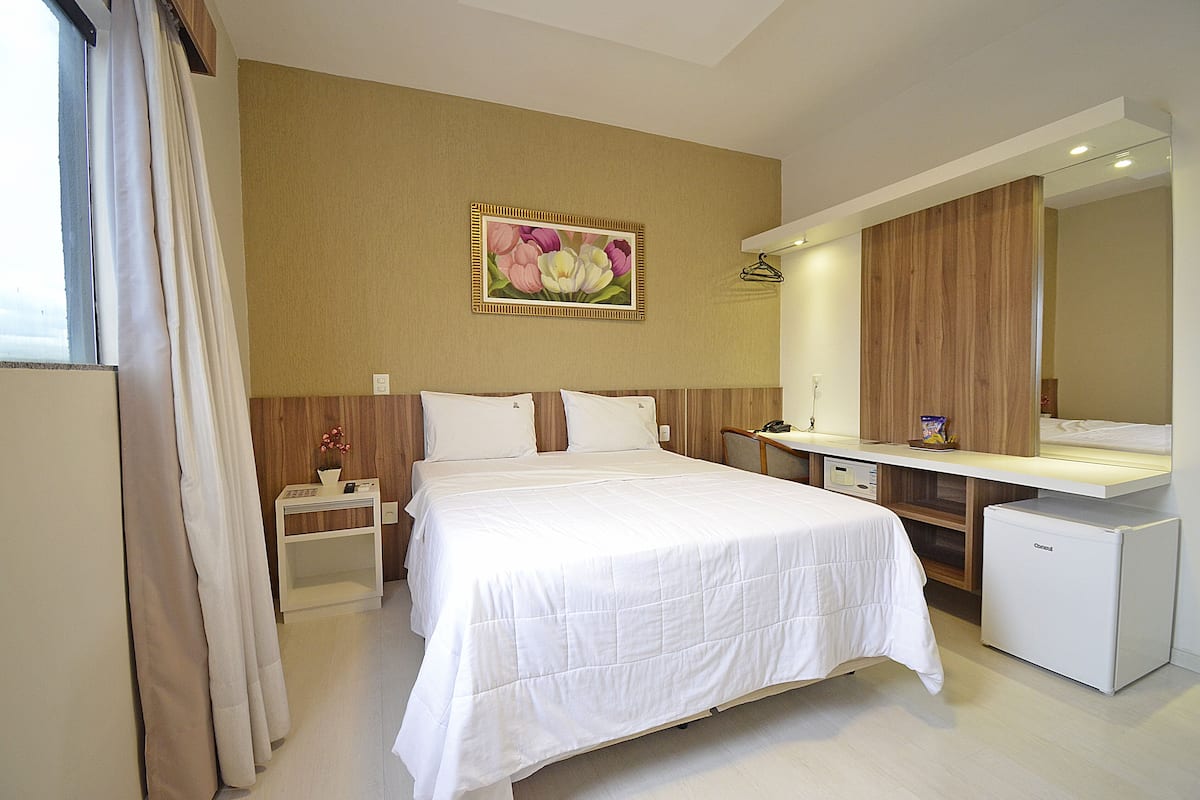 A hotel room with a bed, a nightstand, a desk, a refrigerator, a mirror, and a painting on the wall.