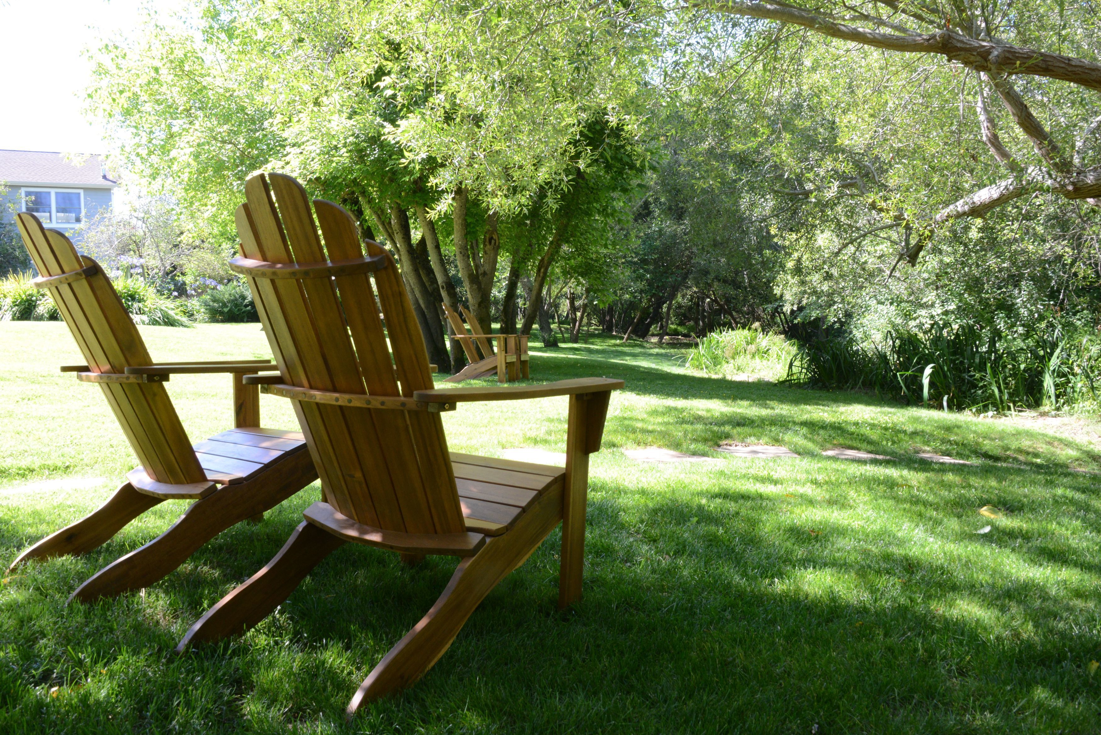 Dos sillas Adirondack de madera en un jardín exuberante con árboles y una casa al fondo.