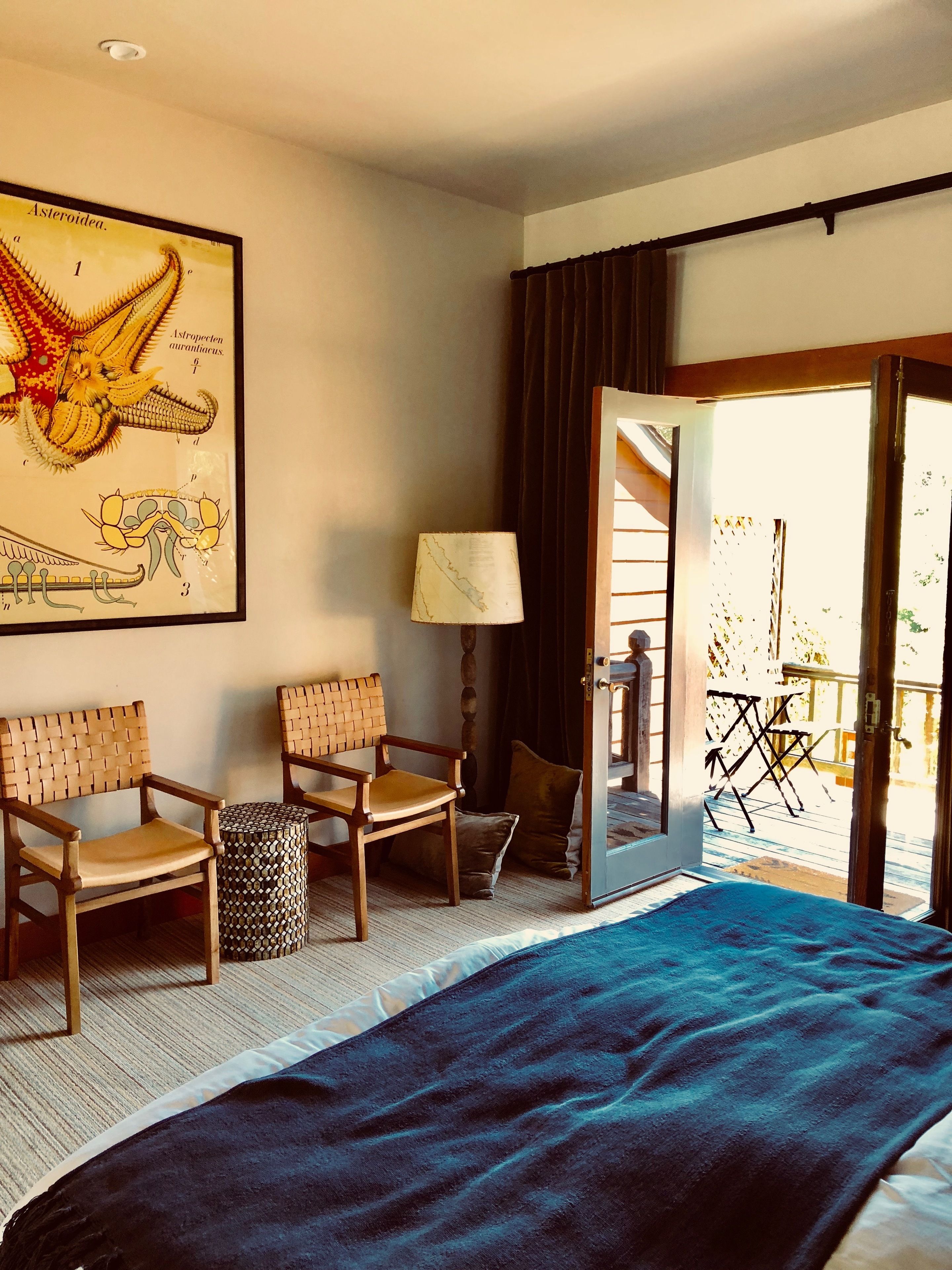 A hotel room with a bed, two wooden chairs, a small table, a lamp, and a large framed illustration on the wall.