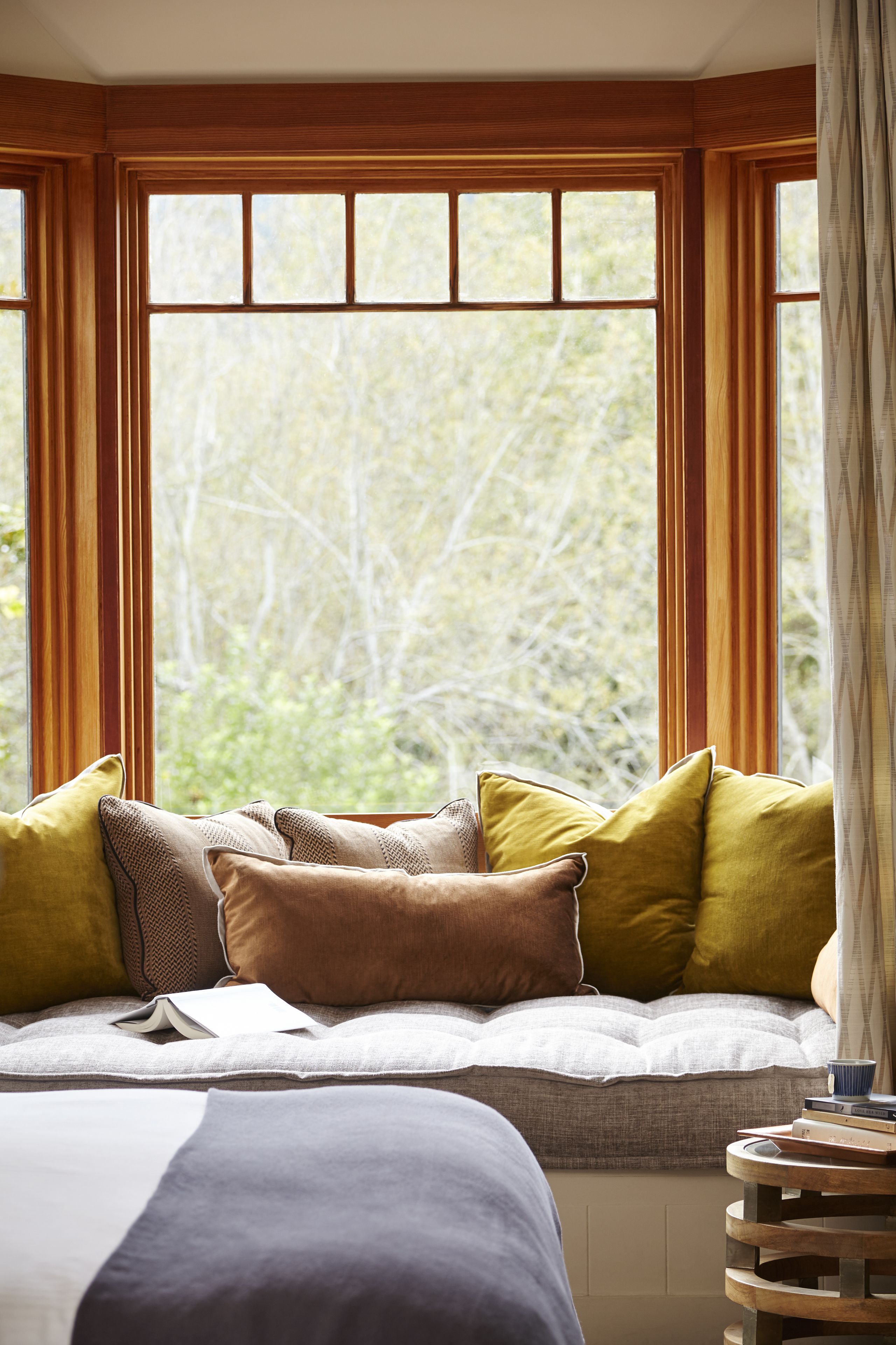 A bedroom with a large window, a bed with multiple pillows, a bedside table with books and a cup, and a curtain on the right.