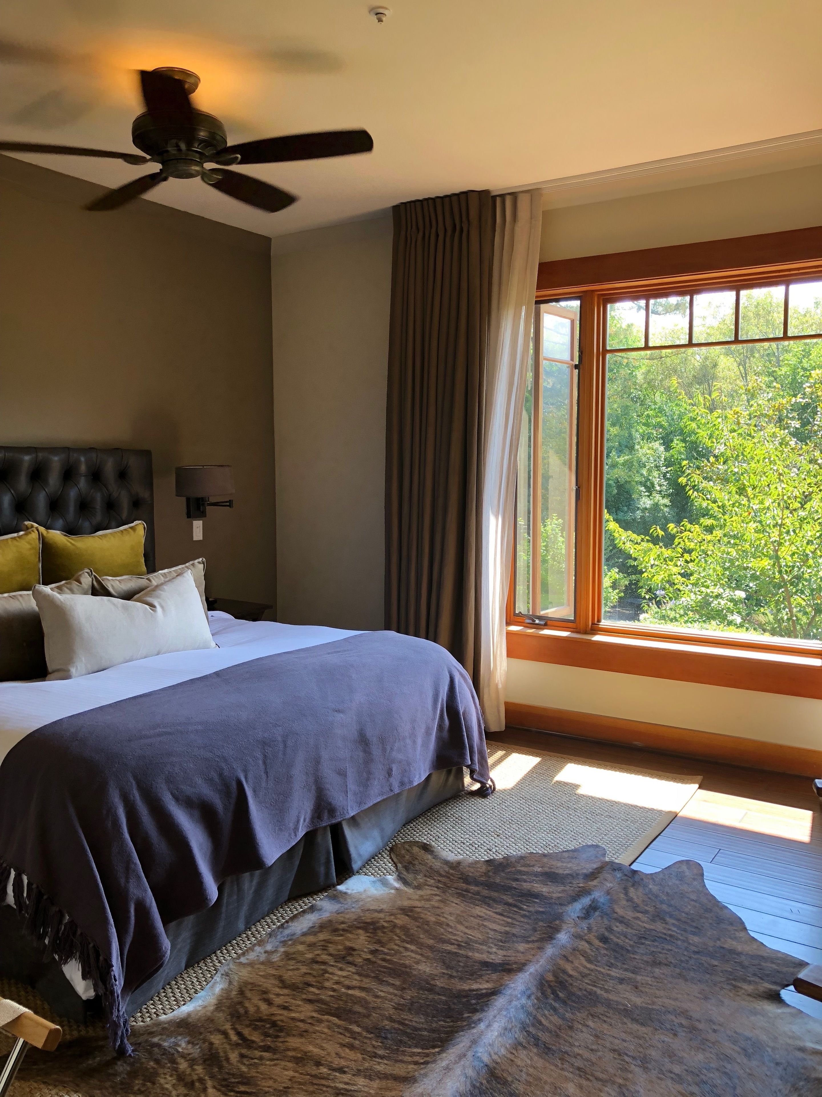 A bedroom with a bed, a ceiling fan, a window with curtains, and a view of trees.