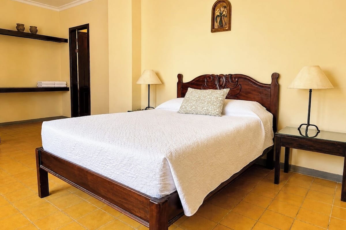 A neatly made bed with a white quilt, two bedside lamps, a wooden headboard, a wall-mounted picture, and a shelf with folded towels.