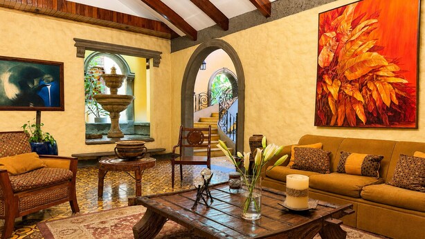 A spacious living room with a wooden coffee table, a sofa, and a TV. There is a large painting on the wall and a fountain in the background. - 1915 Hotel (Alajuela)