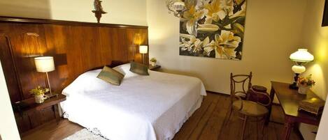 A bedroom with a wooden headboard, a large bed with white bedding, a painting of flowers on the wall, and a desk with a lamp.