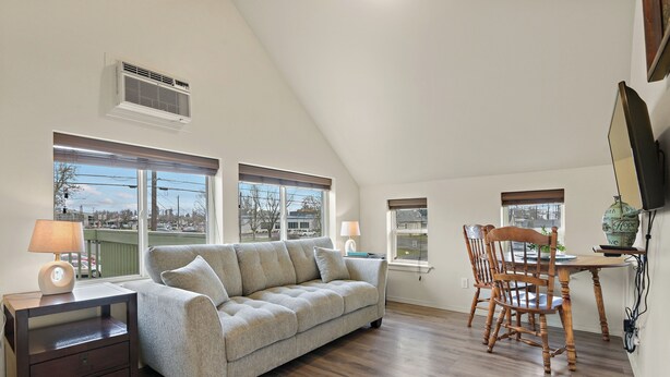 A cozy living room with a sofa, a wooden dining table, and chairs. There are large windows with blinds, a lamp on a side table, and a TV mounted on the wall. - Maple Leaf Manor Suites (Spokane)