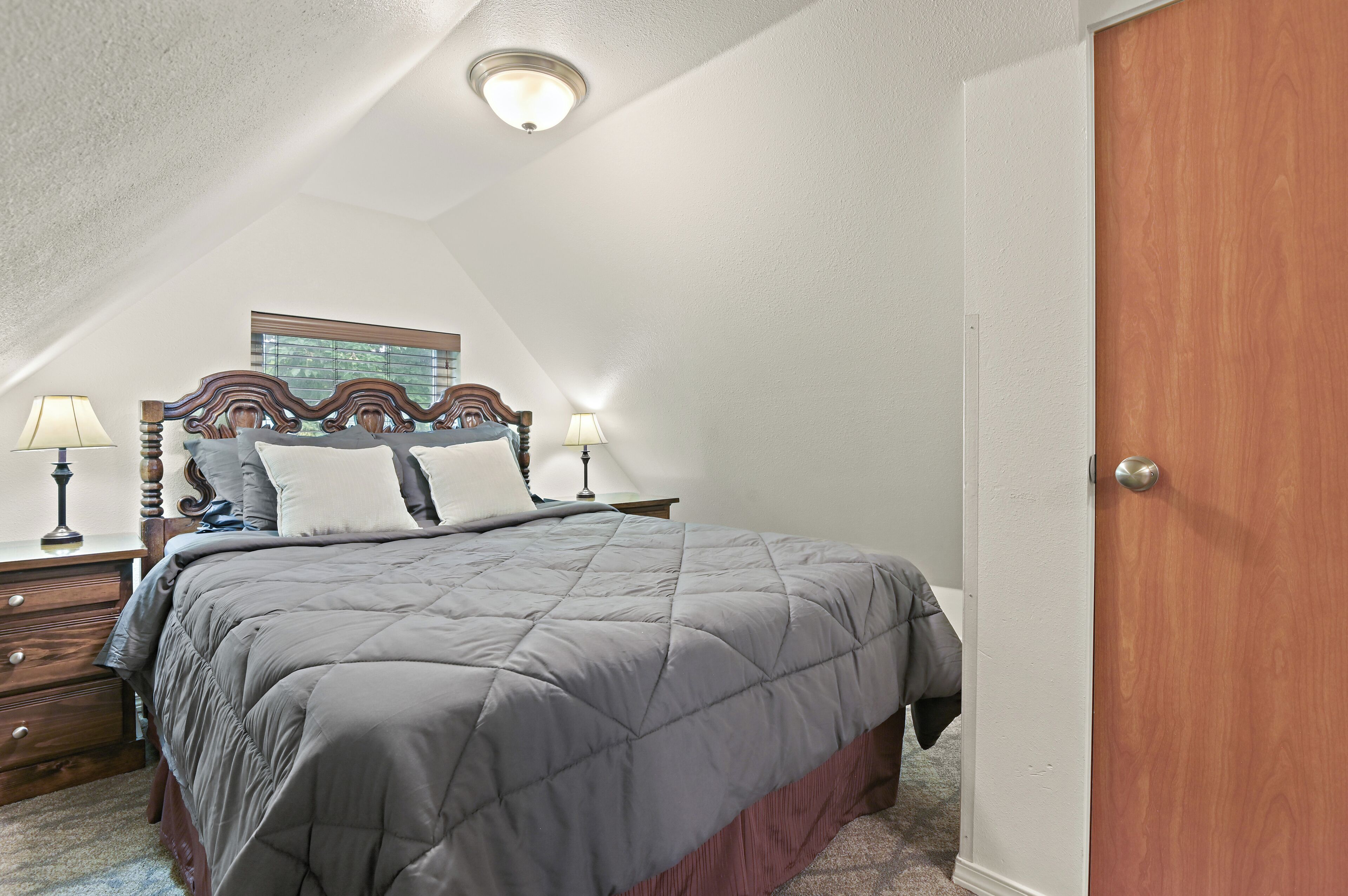 A bedroom with a bed, two bedside lamps, a wooden headboard, a window, and a door.