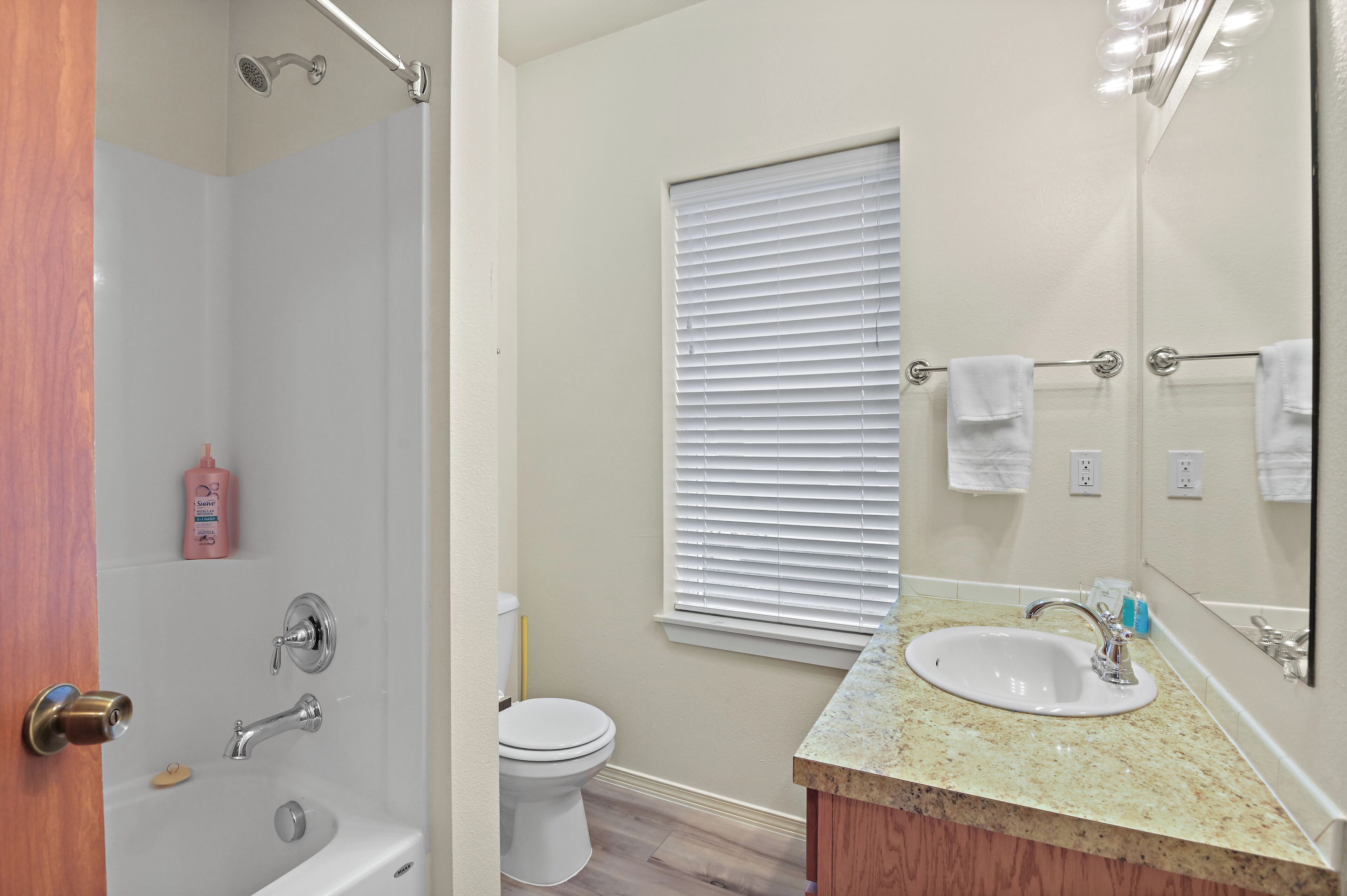 A bathroom with a shower, toilet, sink, and a window with blinds.