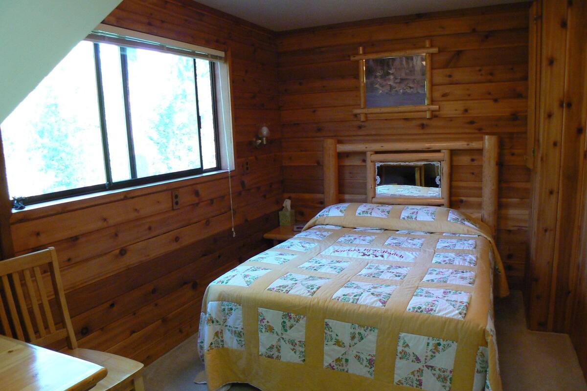 A wooden cabin room with a bed, a chair, a window, and a mirror on the wall.