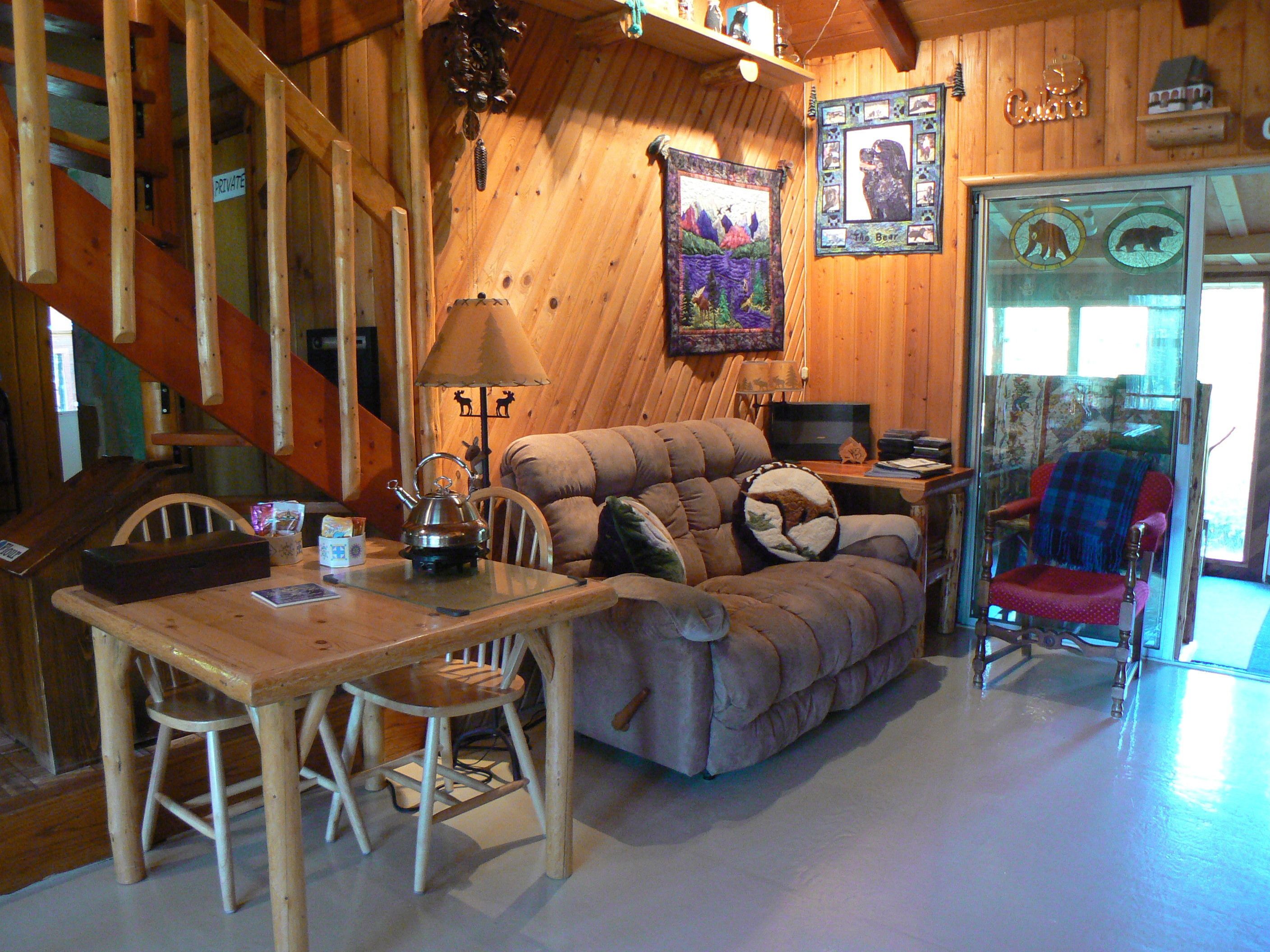 Een gezellig interieur van een chalet met een houten trap, een bank, een eettafel met stoelen en een rode fauteuil.