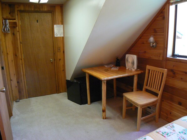 A small, cozy room with a wooden door, a table with a fan, and a chair.