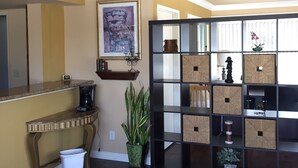 A living room with a dark wood floor, a coffee station, a potted plant, a framed artwork, and a shelving unit with storage boxes. - Fullerton Inn - Near Medieval Times (Fullerton)