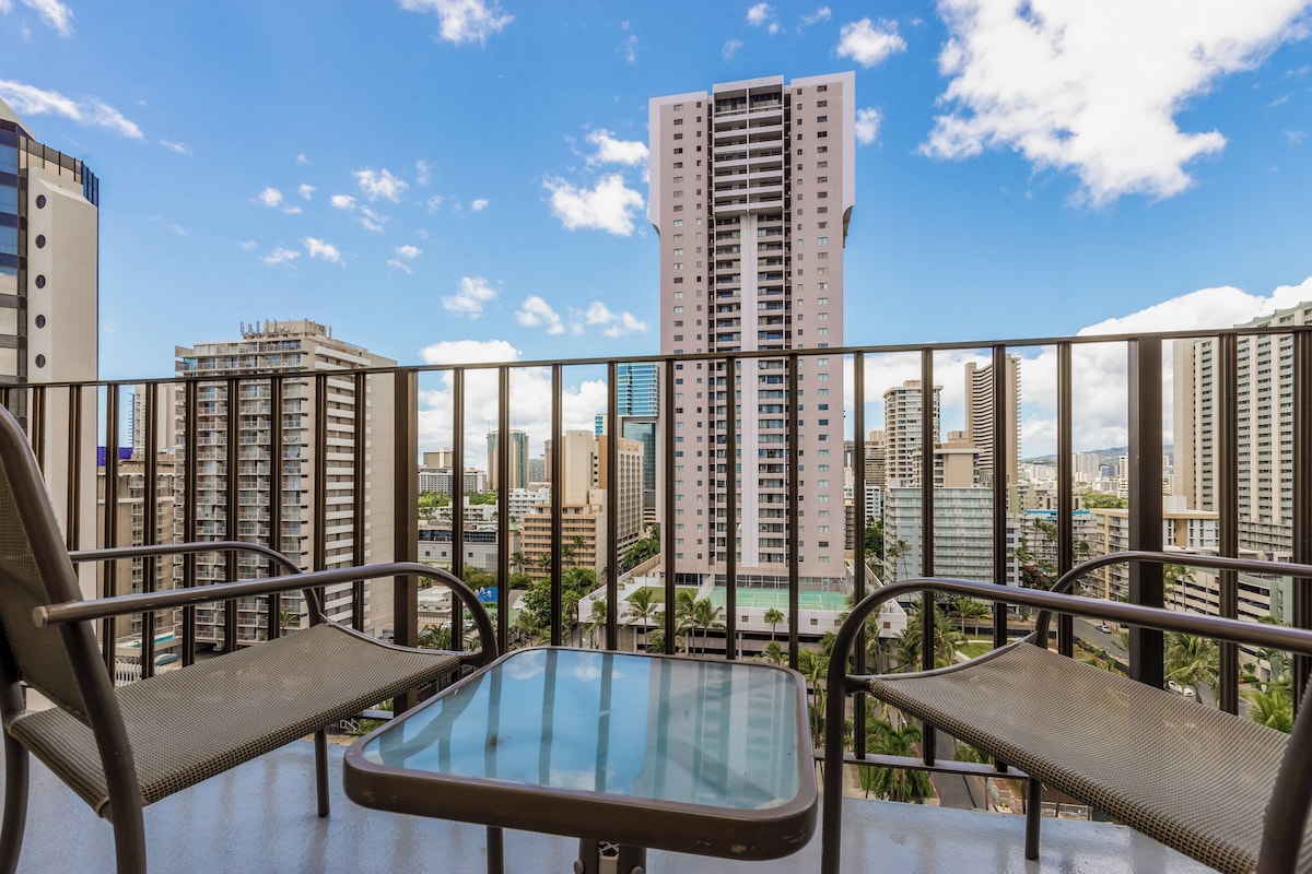 A balcony with two chairs and a small table, overlooking a cityscape with tall buildings and a clear sky.