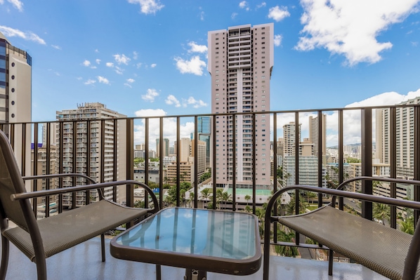 A balcony with two chairs and a small table, overlooking a cityscape with tall buildings and a clear sky.