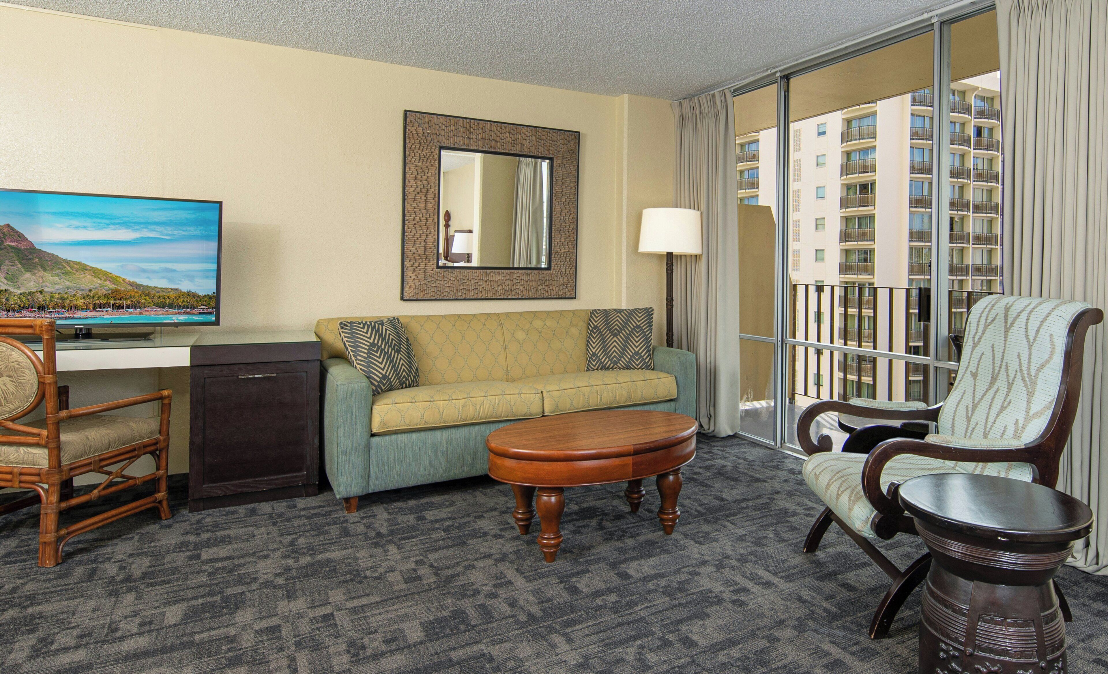 A hotel room with a sofa, armchair, small table, TV, and a large window with a view of buildings.